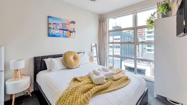 A hotel room with a large bed, a painting on the wall, a window with a city view, and a television on a stand. - Cosy at The Cambridge Apt with Wi-Fi and Netflix (Auckland CBD)