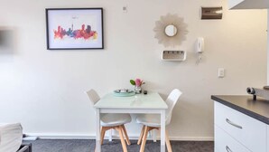A small, modern kitchen with a white table, two chairs, and a framed cityscape picture on the wall. - Cosy at The Cambridge Apt with Wi-Fi and Netflix (Auckland CBD)