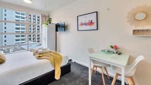 A hotel room with a bed, a desk, a chair, a TV, and a large window with a city view. - Cosy at The Cambridge Apt with Wi-Fi and Netflix (Auckland CBD)