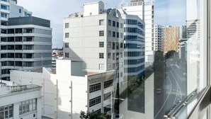 A modern urban street with multi-story buildings, including a prominent white building with a curved facade and a checkered pattern building in the background. - Cosy at The Cambridge Apt with Wi-Fi and Netflix (Auckland CBD)
