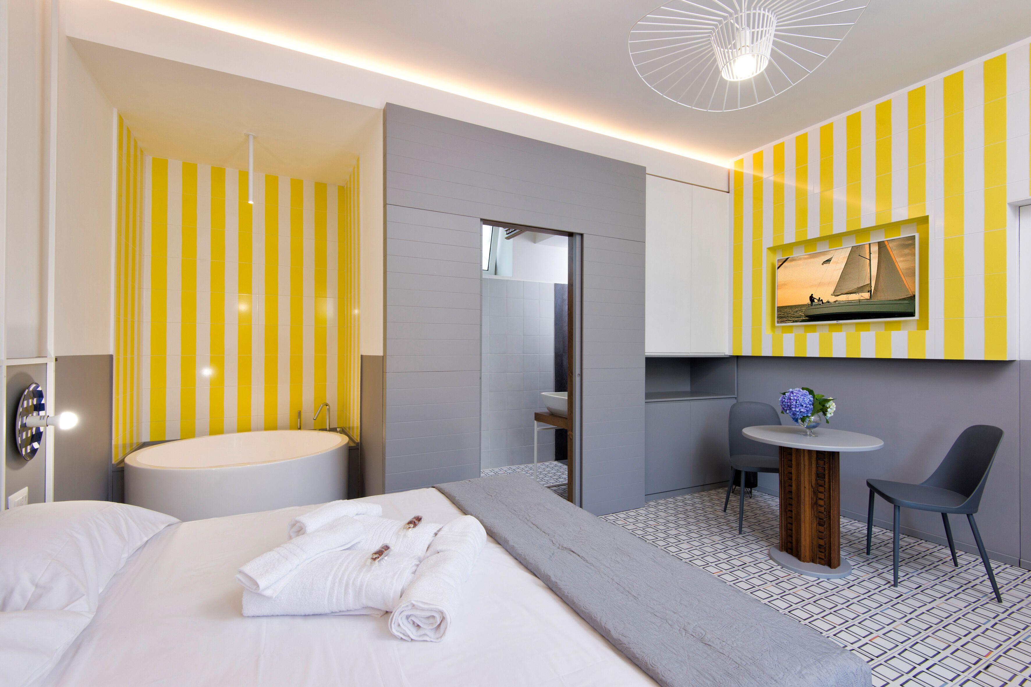 Habitación de hotel moderna con una cama grande, una mesa redonda con dos sillas, una mesita redonda con un florero y una bañera amplia, además de un espejo y una pared a rayas amarillas y blancas.