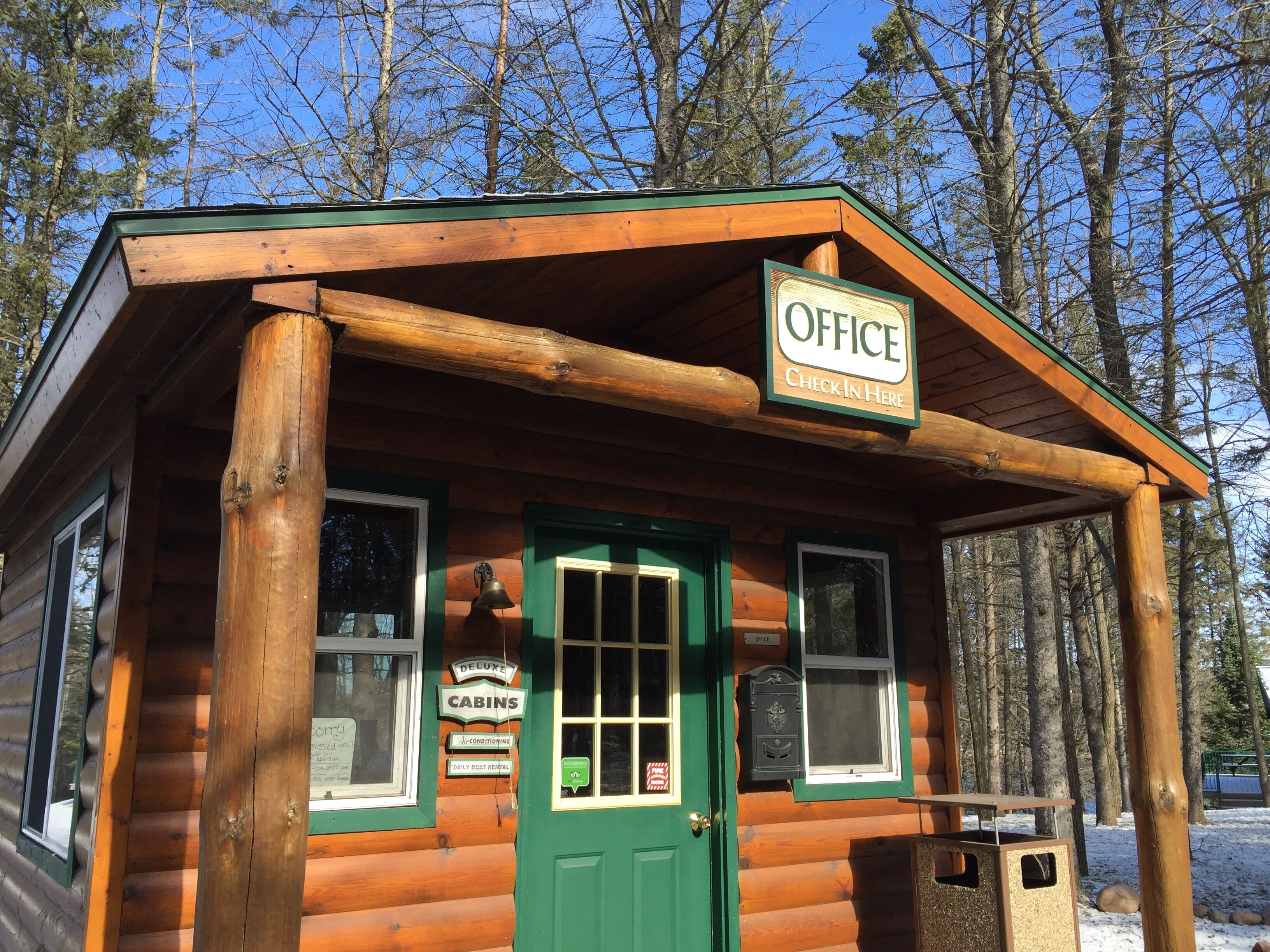 A wooden cabin with a green door, labeled "Office Check In Here," and signs for "Deluxe Cabins" and "Daily Boat Rental."