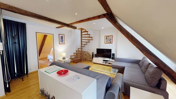 Un salon moderne de style industriel, avec des poutres en bois apparentes, un canapé d’angle gris, un téléviseur à écran plat et un escalier menant à un niveau supérieur.