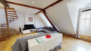 Une pièce de style « loft » avec un escalier en bois, un téléviseur à écran plat, un canapé gris et un meuble blanc orné d’un motif représentant un paysage urbain.