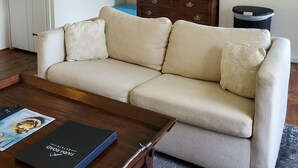 Una sala de estar con un sofá color beige, una mesa de centro de madera, un espejo enmarcado, un microondas, una lámpara y un cubo de basura.