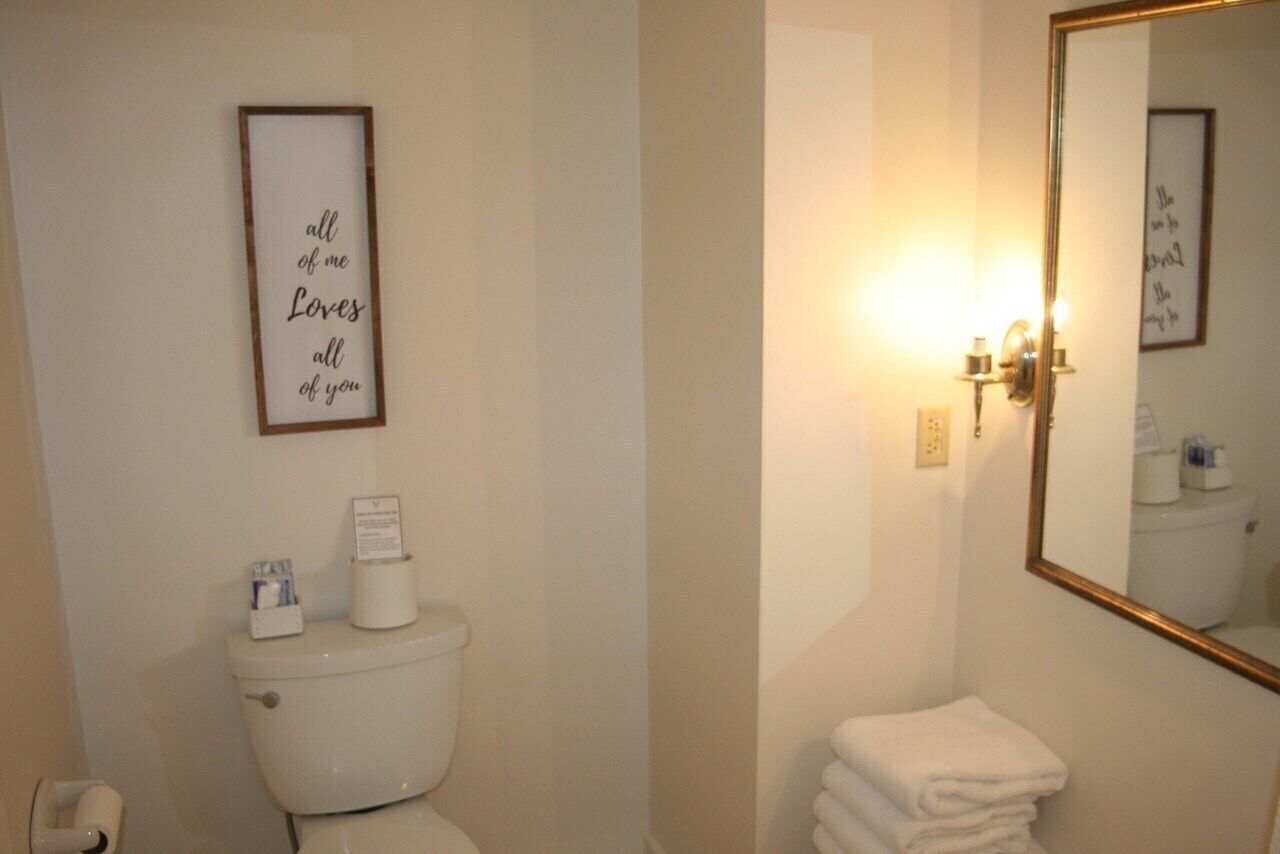 A bathroom with a toilet, a framed sign above it, a wall-mounted light, and a mirror with a gold frame.