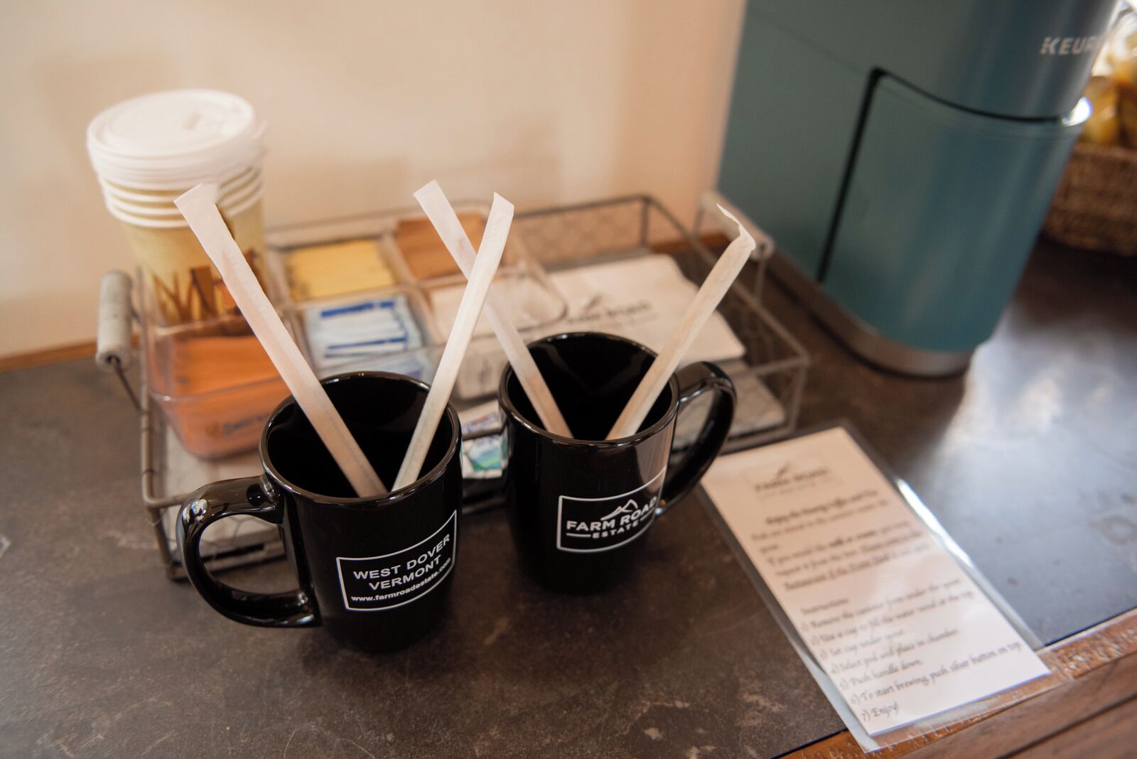 Dos tazas negras con texto blanco y pajitas sobre una bandeja metálica.