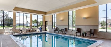 An indoor swimming pool with clear views of the outdoors, surrounded by chairs and tables.