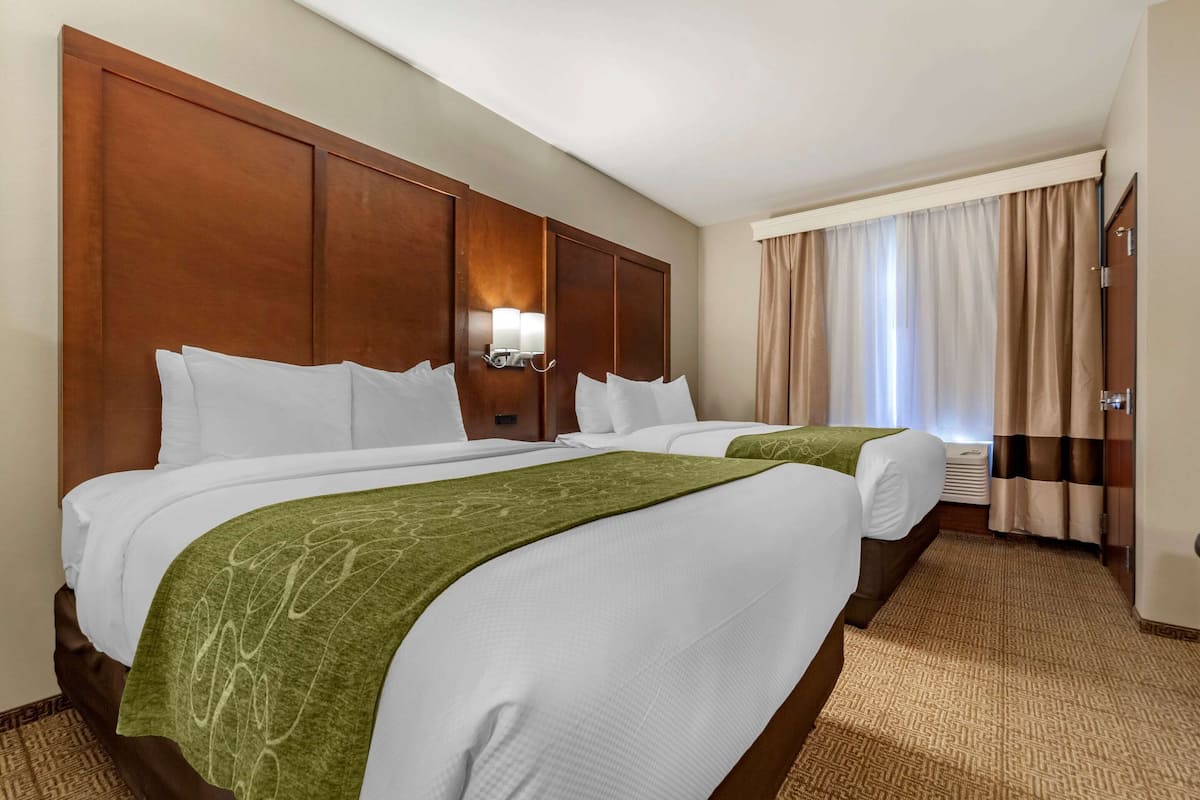 Ett hotellrum med två sängar, en sänggavel i trä, en vägglampa och ett fönster med gardiner.