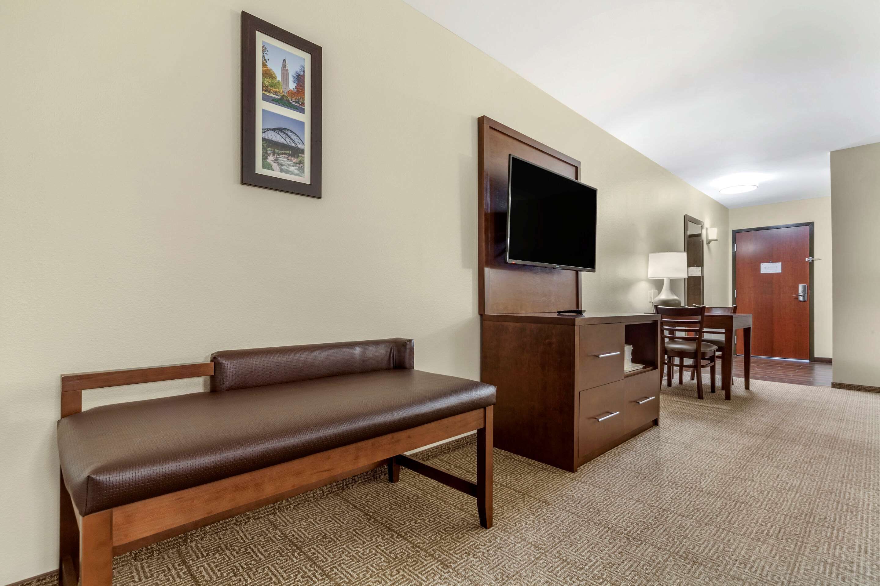 Une chambre d’hôtel avec un banc en bois, un bureau avec une chaise, un téléviseur à écran plat, une lampe et un tableau encadré au mur.