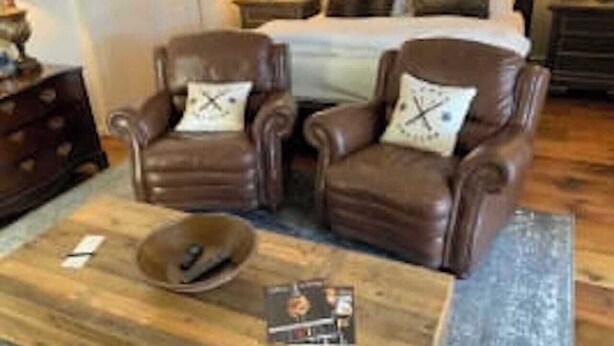 Ein Zimmer mit zwei braunen Ledersesseln, einem Holztisch mit Schale und Zeitschrift sowie einem Bett mit Kissen im Hintergrund.