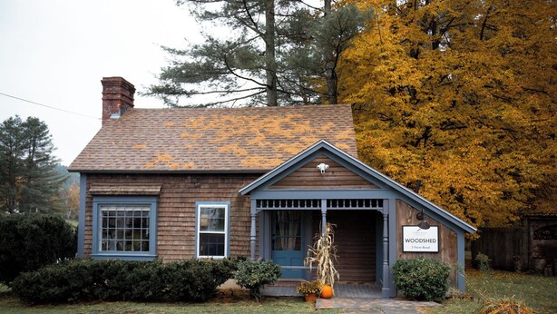 A small, two-story wooden house with a porch, a chimney, and a sign indicating it is a woodshedd.