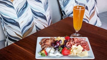 A charcuterie board with various meats, cheeses, and fruits on a rectangular plate, accompanied by a glass of beer, set on a wooden table with patterned cushions in the background.
