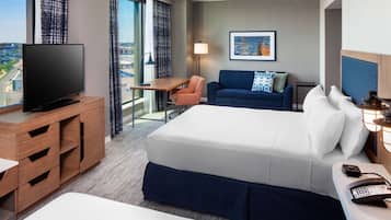 A hotel room with a large bed, a desk with a TV, a chair, a sofa, and a city view.