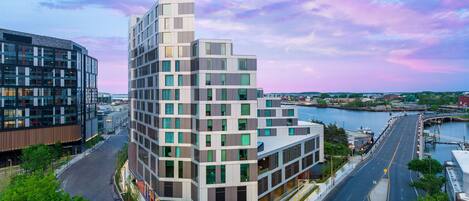 A modern building with a glass facade, located near a waterfront with a bridge and a road.