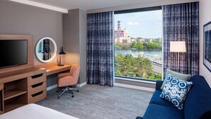 Une chambre d’hôtel comprenant un lit, un bureau avec une télévision, une chaise, un canapé avec un coussin à motifs, et donnant sur un plan d’eau et des bâtiments.