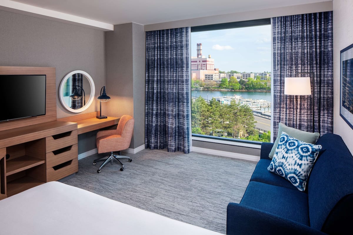 A hotel room with a bed, a desk with a TV, a chair, a sofa with a patterned pillow, and a view of a body of water and buildings.