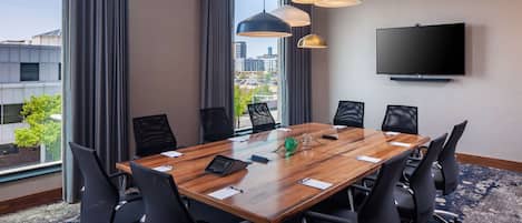 Un salón de conferencias con una amplia mesa de madera, sillas negras, un televisor de pantalla plana y vistas a la ciudad a través de grandes ventanales.