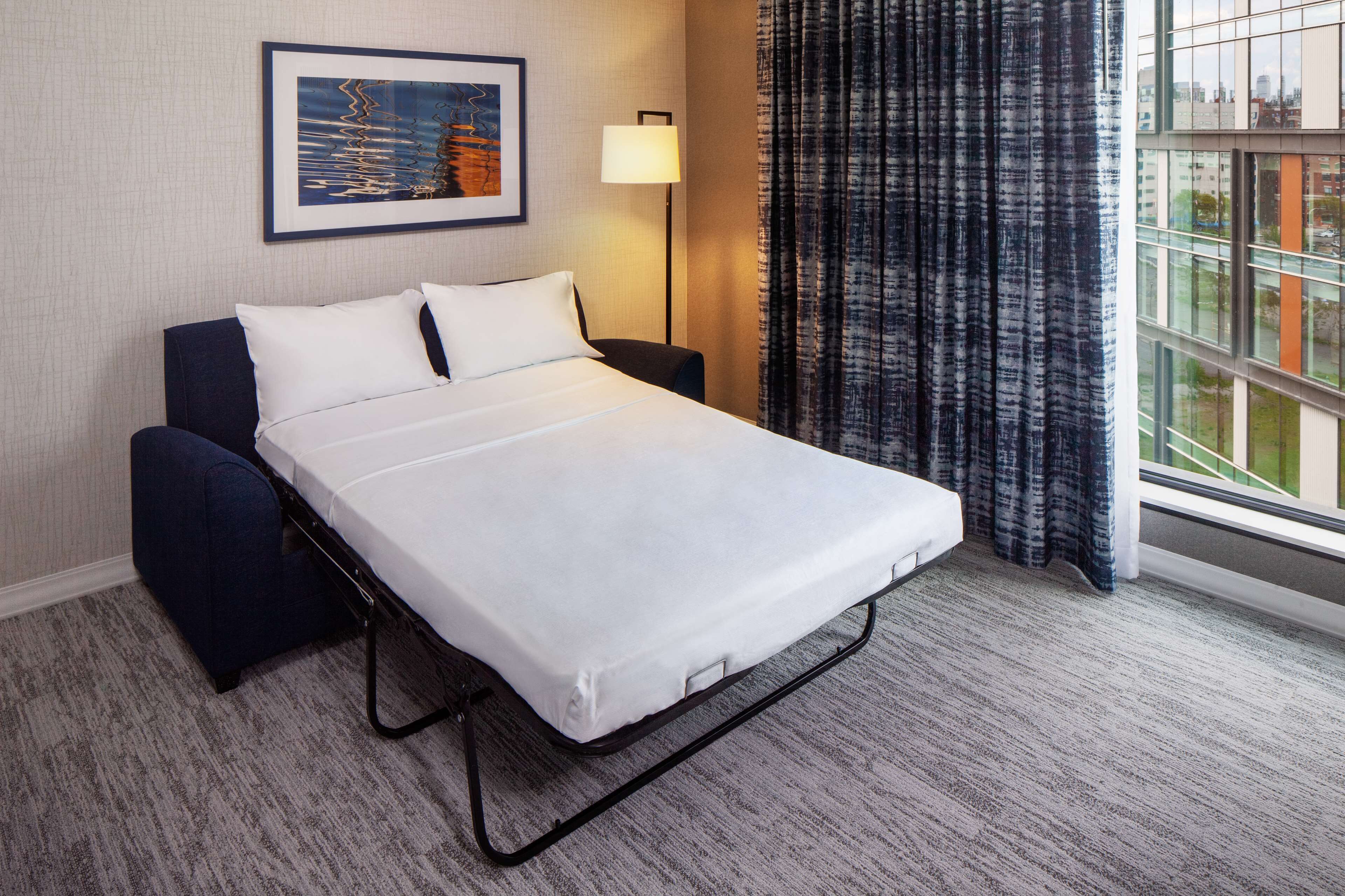 Une chambre d’hôtel avec un lit simple, une table de chevet, une lampe, une fenêtre avec des rideaux et un tableau encadré au mur.