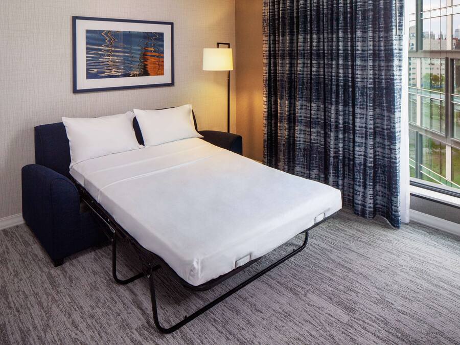 Habitación de hotel con una cama individual, una mesita de noche, una lámpara, una ventana con cortinas y un cuadro en la pared.
