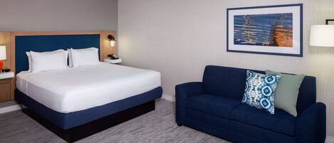 Una habitación de hotel con una cama grande, un sofá azul, un cuadro enmarcado en la pared y una mesita de noche con una lámpara.