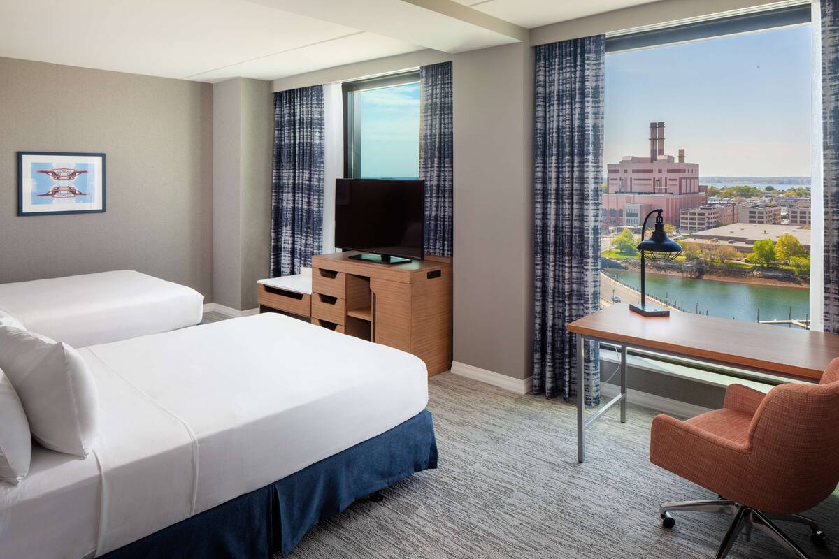 A hotel room with two beds, a desk, a TV, and a view of a cityscape.