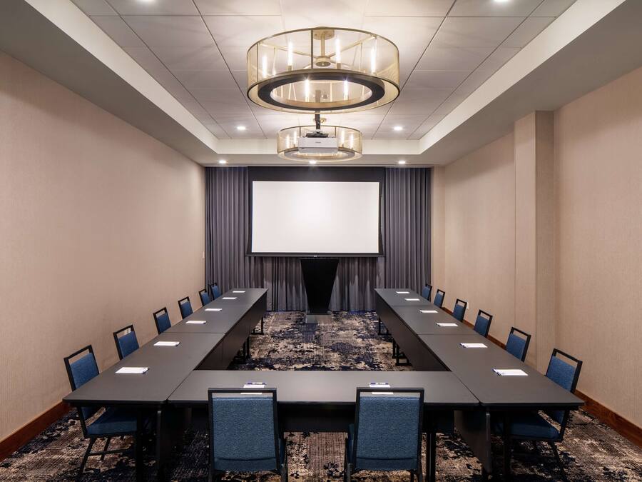 Een vergaderzaal met een lange tafel, stoelen, een projectiescherm en een vloerbedekking.