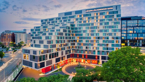 Un hôtel moderne, reconnaissable à son grand panneau « Hampton Inn », composé d’un bâtiment à plusieurs étages avec une cour intérieure et un espace paysager.