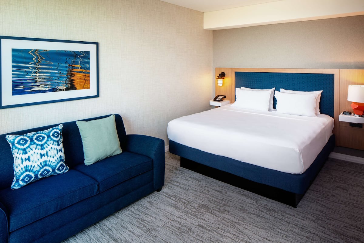 A hotel room with a large bed, a blue sofa, a framed picture on the wall, and a nightstand with a lamp.