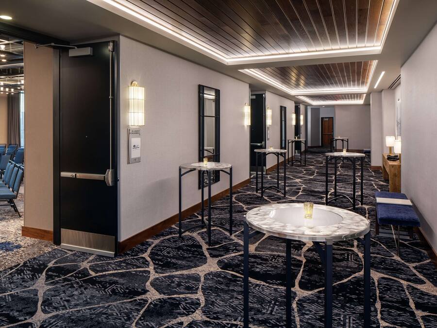 Un couloir d’hôtel avec un tapis à motifs, un coin salon avec des chaises bleues et une table ronde avec un plateau en verre.