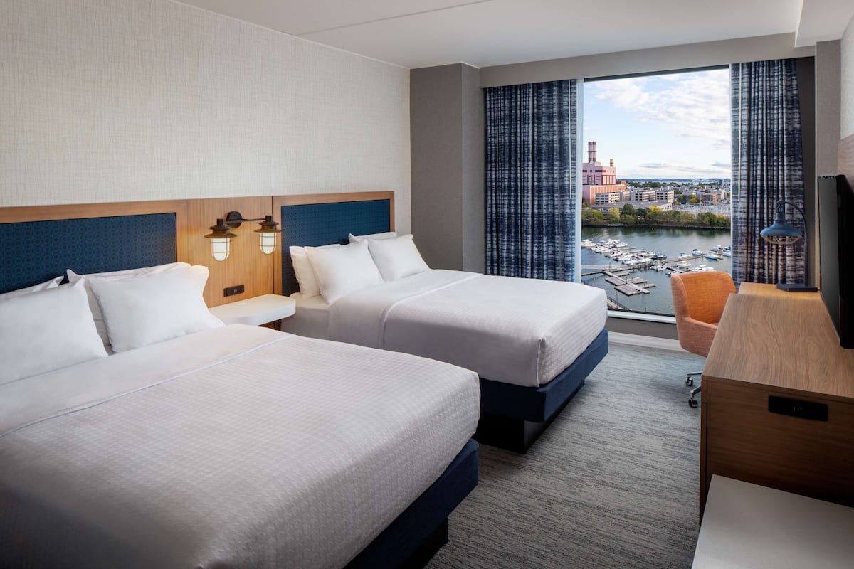 A hotel room with two beds, a desk, a chair, and a large window with a view of a marina and buildings.