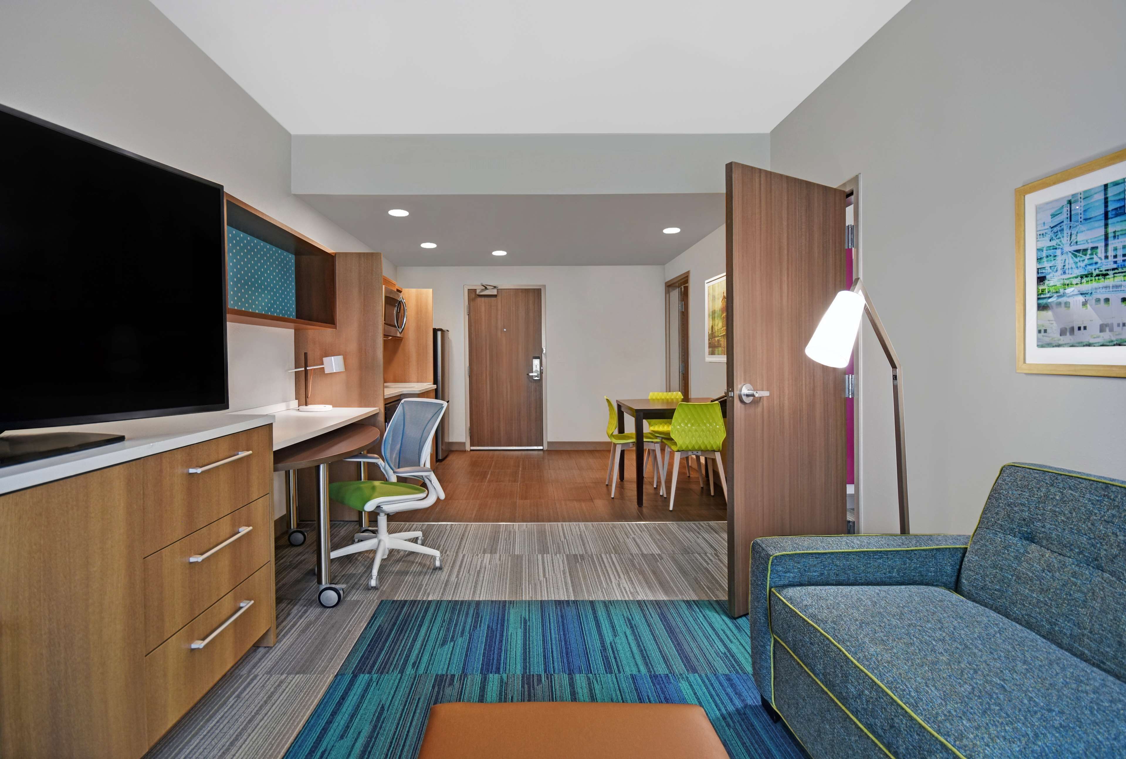 Una habitación de hotel moderna con una televisión de pantalla plana, armarios de madera, un escritorio con silla, una zona de comedor con sillas, un sofá y una lámpara de pie.