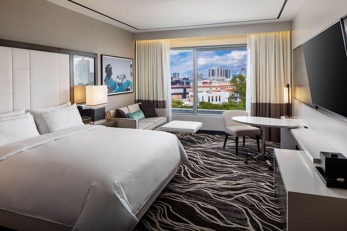 A modern hotel room with a large bed, a desk, a chair, a small table, and a large window offering a city view.