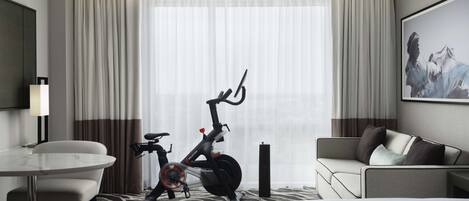 A hotel room with a bed, a desk, a chair, a sofa, a bicycle, and a large window with curtains.