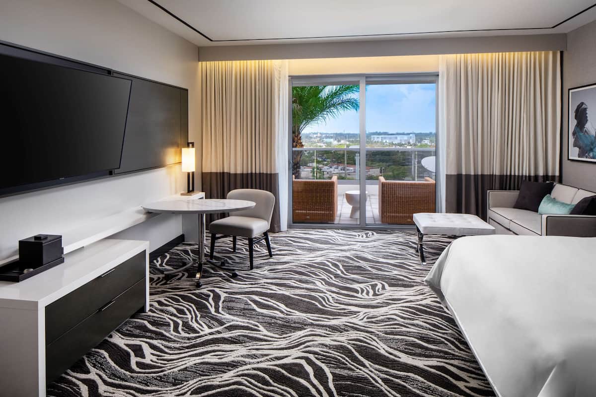 A hotel room with a large bed, a TV mounted on the wall, a small round table, two chairs, a sofa, and a balcony with a view of the city.