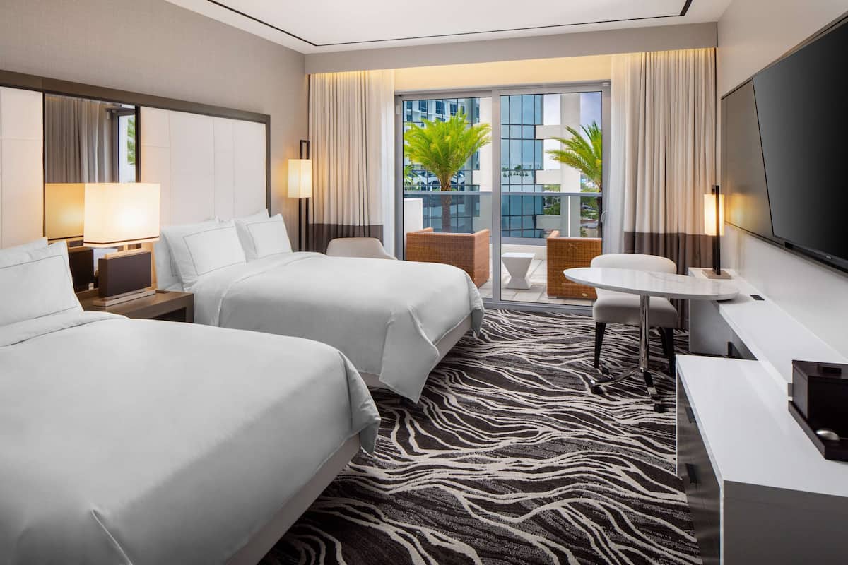 A hotel room with two beds, a desk, a chair, a TV, and a balcony with a view of buildings and a palm tree.