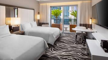 A hotel room with two beds, a desk, a chair, a TV, and a balcony with a view of buildings and a palm tree.