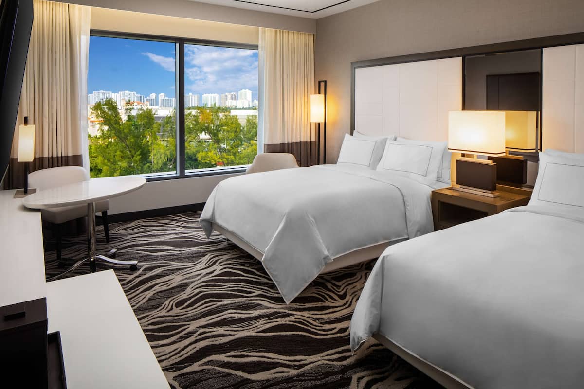 A hotel room with two beds, a desk, and a view of a cityscape.