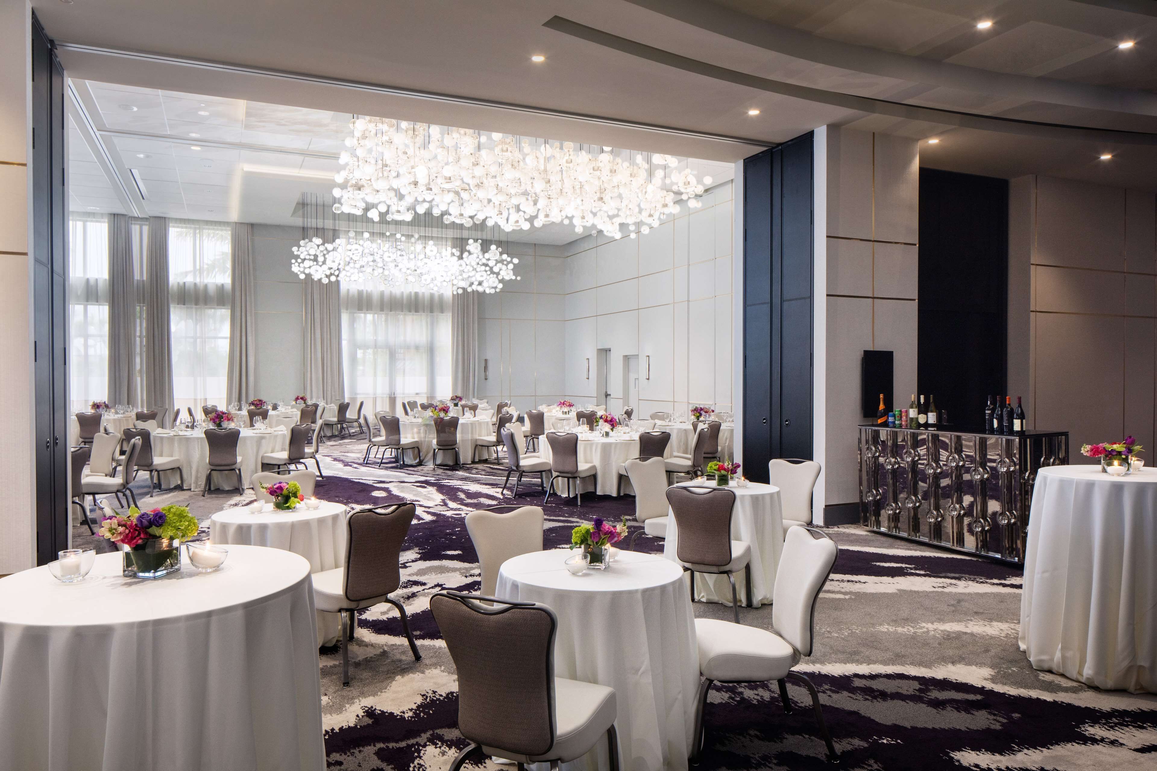 A spacious banquet hall with round tables, white tablecloths, and modern chandeliers.