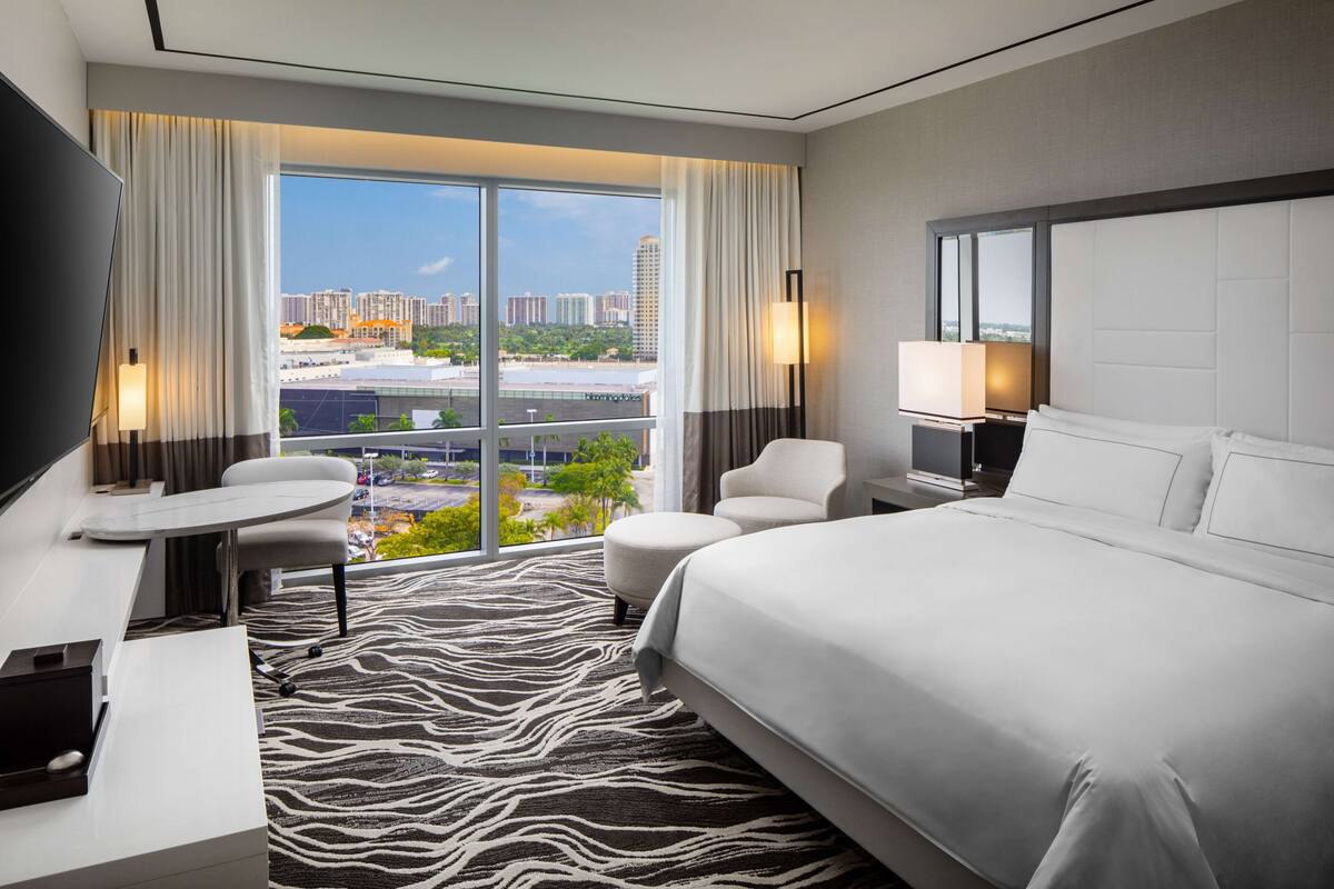 A modern hotel room with a large bed, a desk, a chair, and a TV. There is a view of a cityscape through large windows.