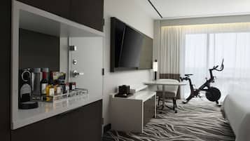 A hotel room with a minibar, a flat-screen TV, a desk with a chair, an exercise bike, and a large window with curtains.