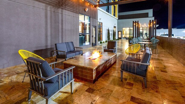 An outdoor seating area with fire pits, chairs, and tables, surrounded by a modern building with large windows and a covered walkway.