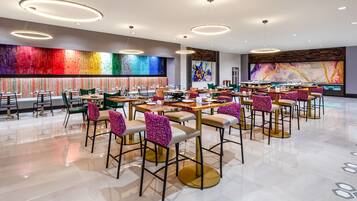 Un área de comedor moderna con varias mesas y sillas, que presenta un mural de colores y lámparas contemporáneas.