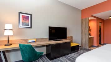A hotel room with a bed, a desk with a TV, a green chair, and a framed picture on the wall.