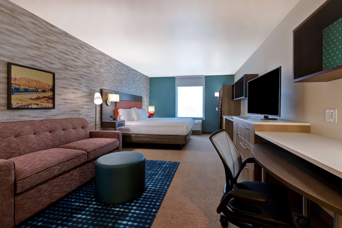 A hotel room with a bed, a sofa, a desk, a chair, a TV, and a painting on the wall.