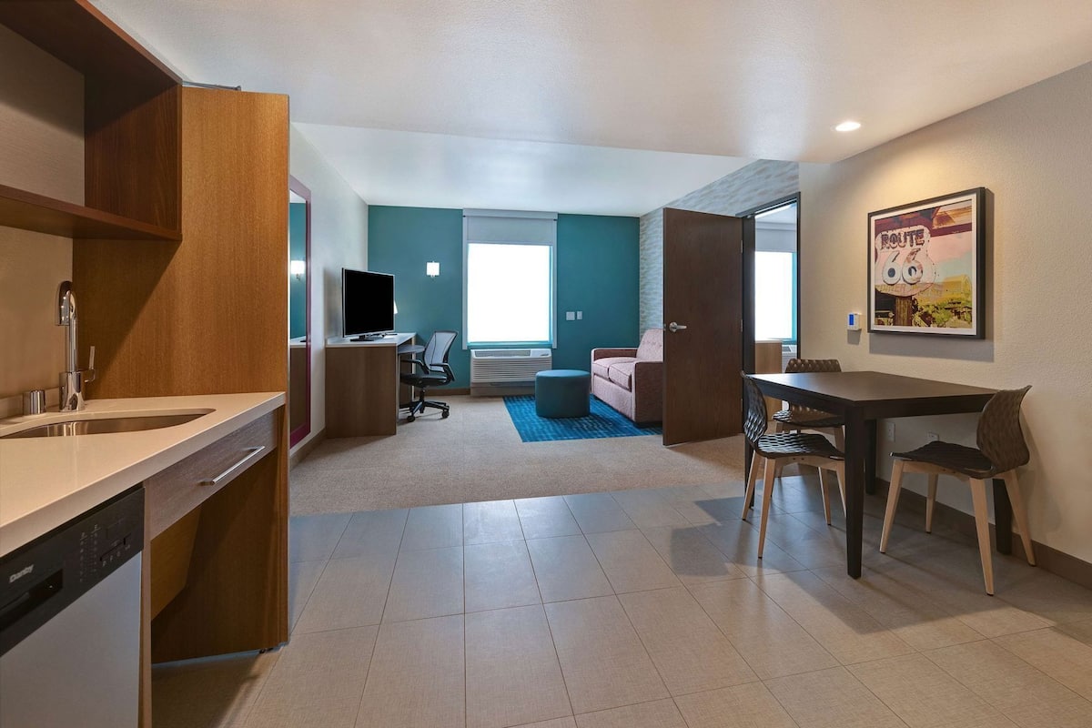 A modern hotel room with a kitchenette, a dining area, a sofa, and a TV.