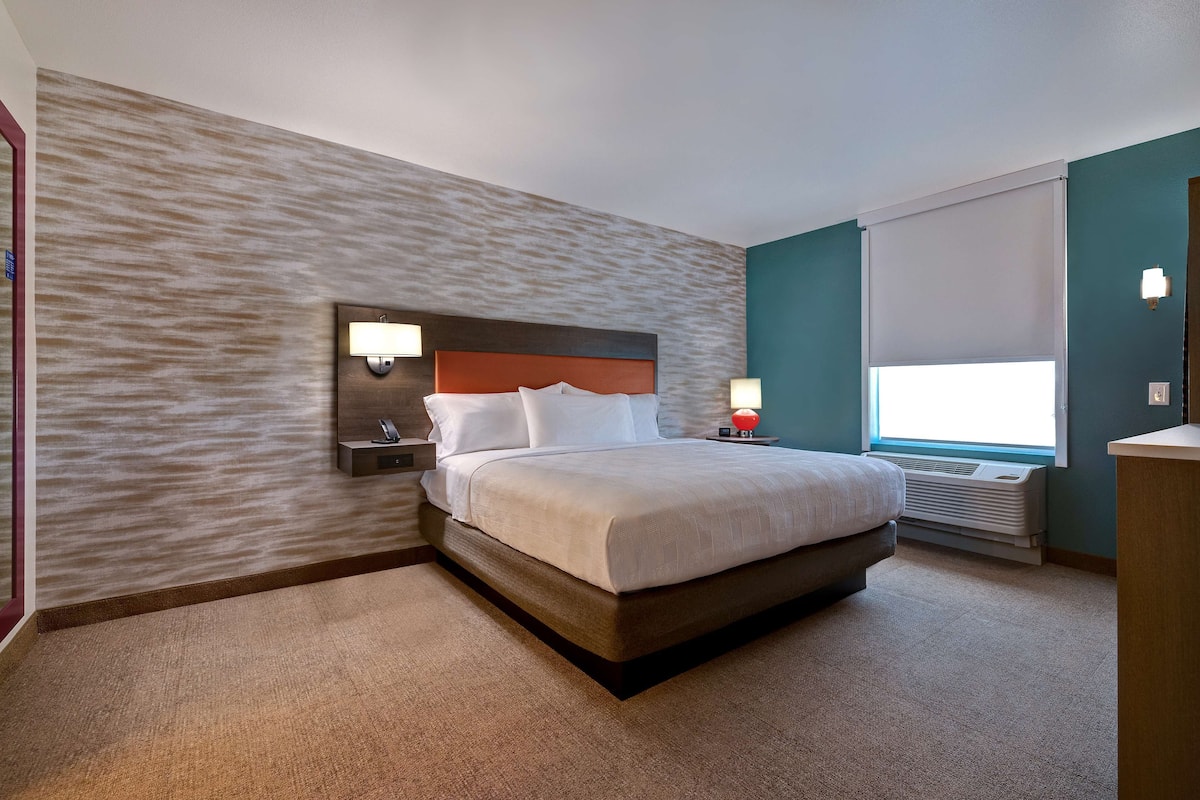 A hotel room with a large bed, two bedside tables with lamps, a wall-mounted heater, and a window with blinds.