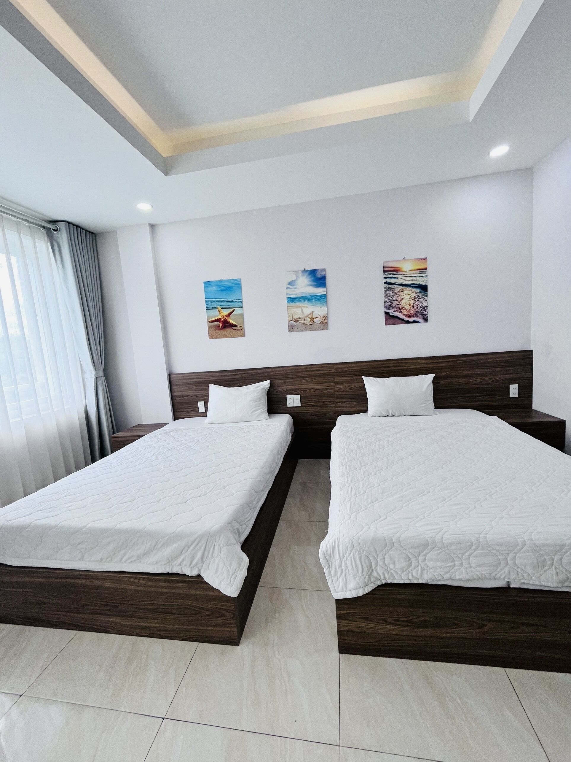 Une chambre d’hôtel avec deux lits, une grande fenêtre avec des rideaux et trois œuvres d’art murales représentant des scènes de plage.