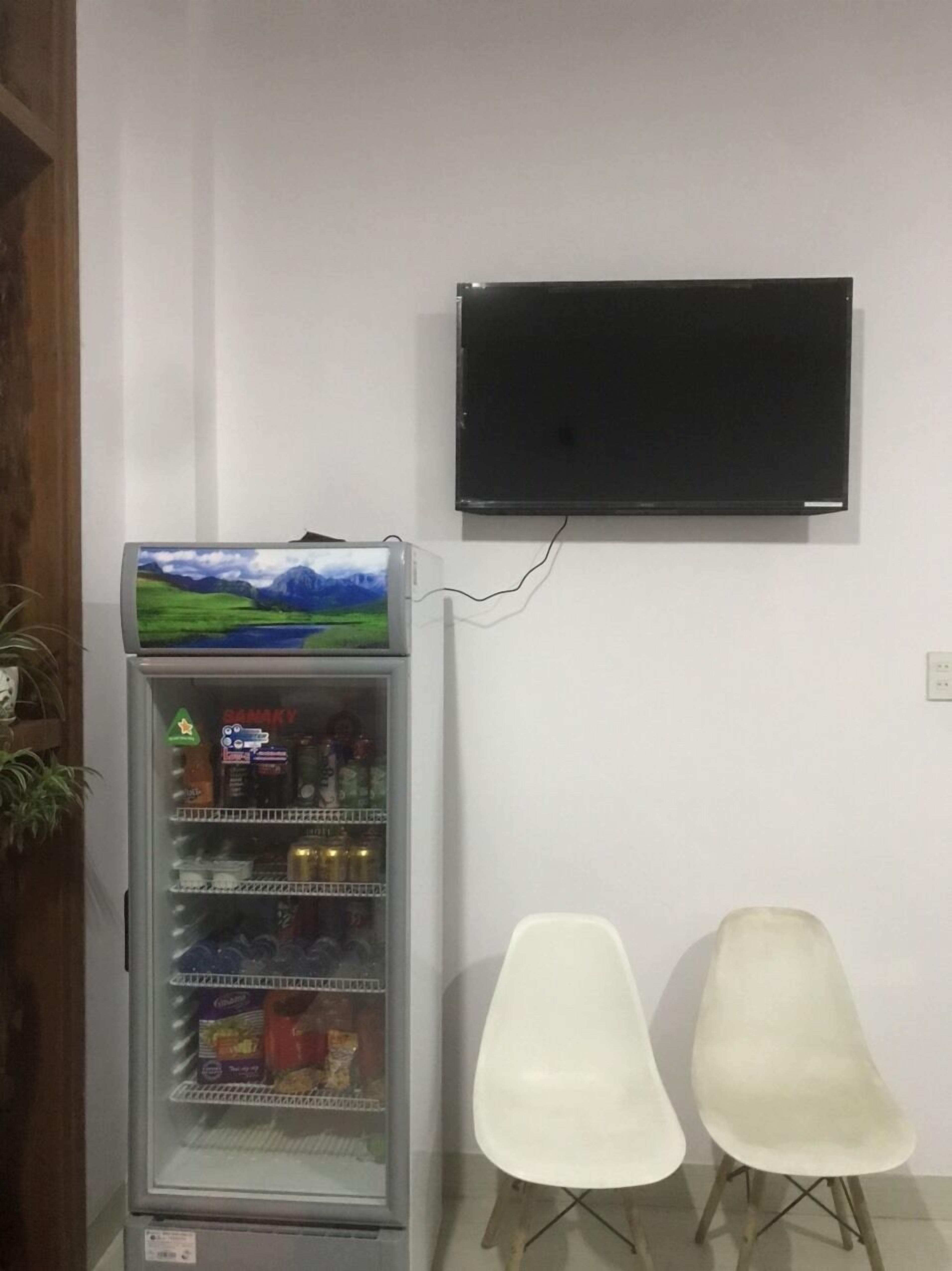 A room with a refrigerator stocked with beverages, two white chairs, and a mounted television on the wall.