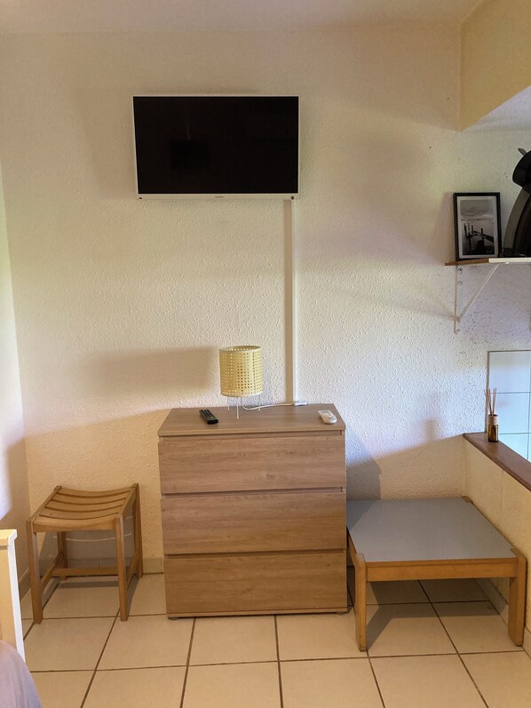 A room with a wooden dresser, a small stool, a wooden table, a mounted television, and a framed picture on the wall.